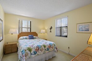 A bedroom with a bed, two bedside tables, a nightstand, a lamp, a window with blinds, and a wall-mounted picture. - 💸 Ground-Floor 1BR Condo — Simple Comfort & Great Value at Gulf Winds (ST PETE BEACH)