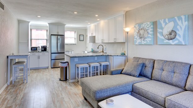 A modern kitchen with white cabinets, a central island with stools, and a grey sofa in the living area.