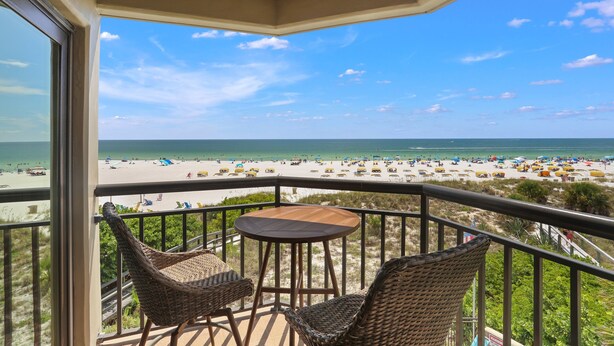 A balcony with wicker chairs and a small table, overlooking a beach with umbrellas and people.