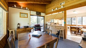 Interior de una cabaña de madera con mesa, sillas, una cocina y un ventanal grande.