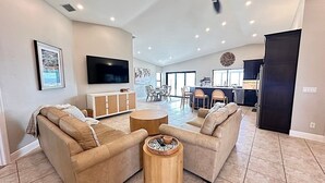 A spacious living room with a large TV, beige sofas, a wooden coffee table, and a dining area in the background. - The River View 1008 - SE Cape Coral Riverfront Luxury Pool Home Pool Table and more (Cape Coral)