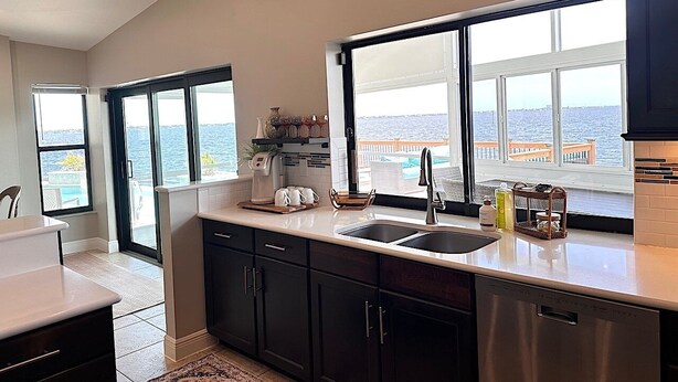 A modern kitchen with a large window overlooking a pool and the sea. It includes a sink, a coffee maker, and various kitchen items on the counter. - The River View 1008 - SE Cape Coral Riverfront Luxury Pool Home Pool Table and more (Cape Coral)