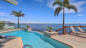 A swimming pool with a view of the ocean, palm trees, and a deck with lounge chairs. - The River View 1008 - SE Cape Coral Riverfront Luxury Pool Home Pool Table and more (Cape Coral)