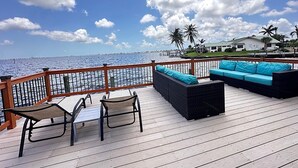 A wooden deck with outdoor furniture, a view of the ocean, and palm trees in the background. - The River View 1008 - SE Cape Coral Riverfront Luxury Pool Home Pool Table and more (Cape Coral)