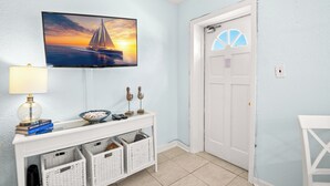 Una habitación con televisión montada en la pared, una mesita consola blanca con cestas, una lámpara y una puerta con ventanal.