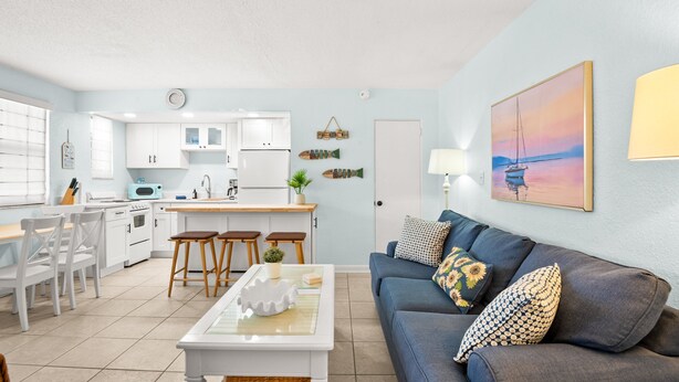 Una cocina moderna con armarios blancos, un comedor con sillas blancas y una sala de estar con un sofá azul y cojines decorativos.
