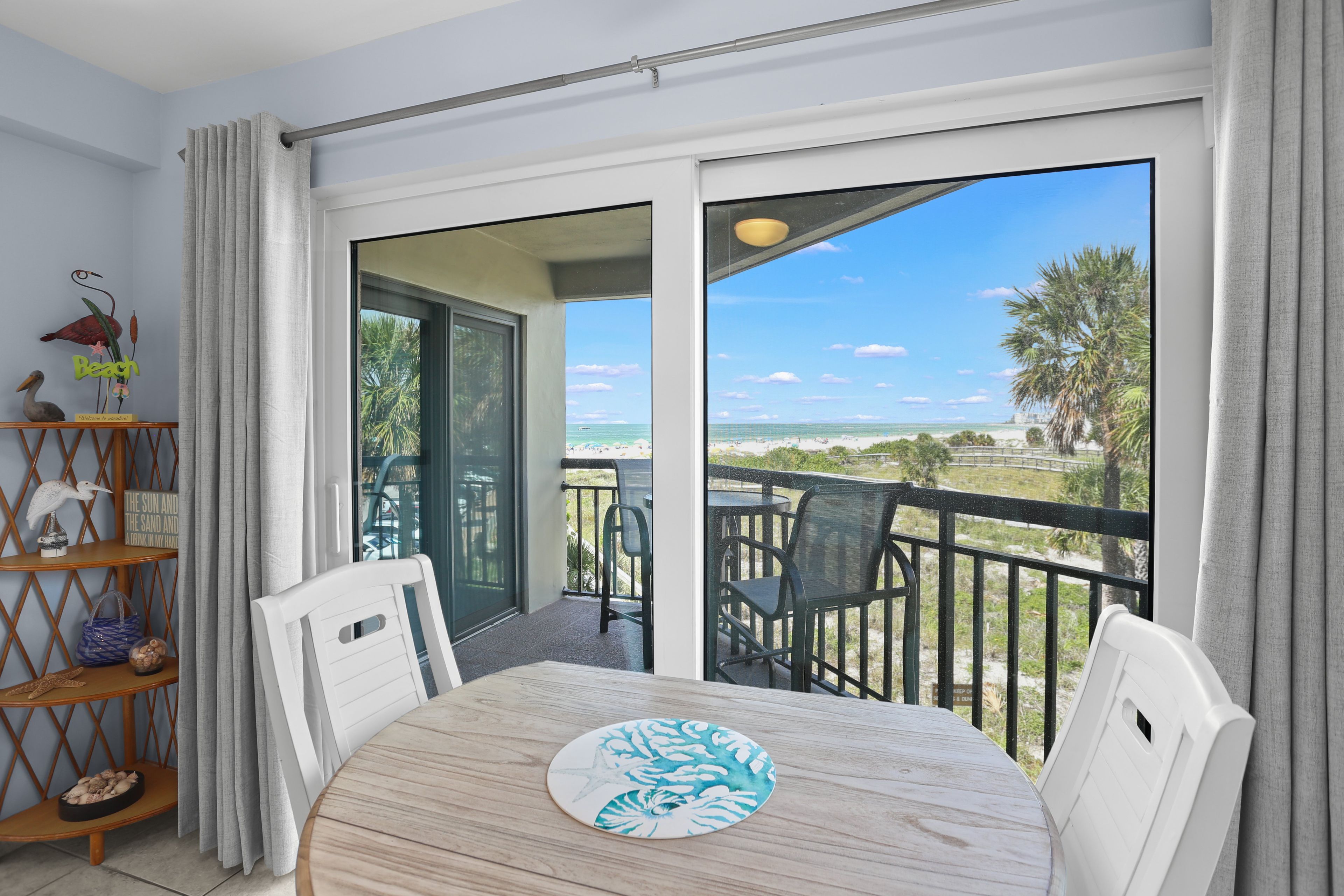 A balcony with a view of the beach, a table with chairs, and a decorative shelf with items.