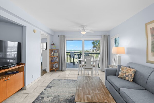 A living room with a television, sofa, coffee table, and a view of the beach through large windows.