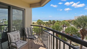 Ein Balkon mit zwei gepolsterten Stühlen, Blick auf den Strand, Palmen und einen klaren Himmel.