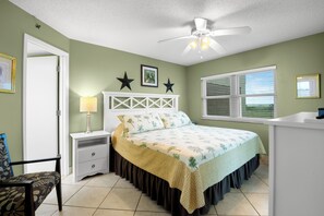 Une chambre à coucher comprenant un lit, des tables de chevet, une chaise, un bureau, un ventilateur de plafond et une fenêtre avec des stores.