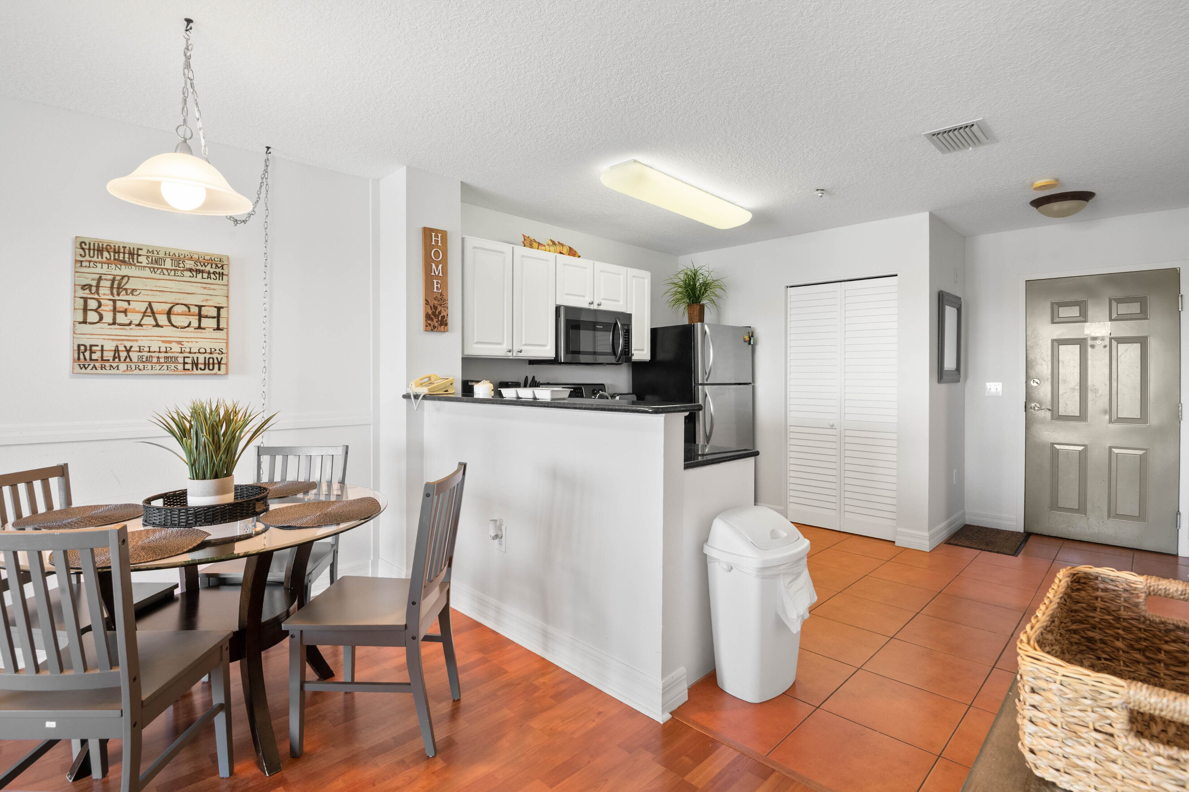 A compact kitchen with white cabinets, a black refrigerator, and a microwave. There's a dining table with chairs and a hanging light fixture.