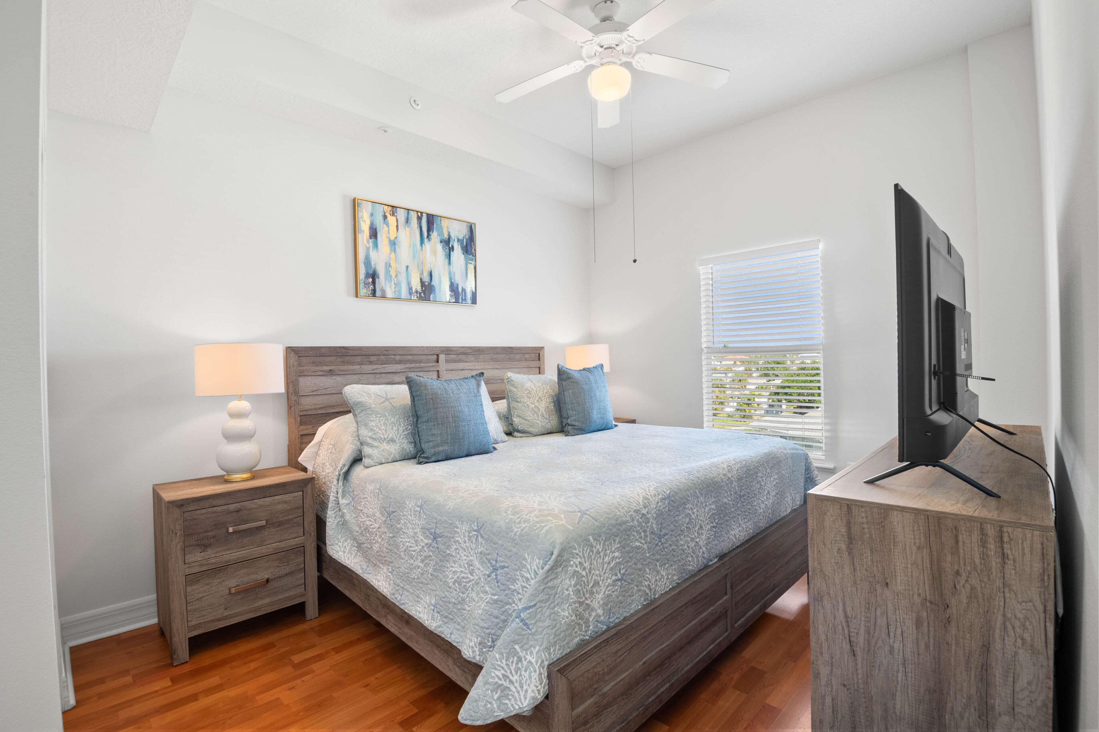 Une chambre à coucher avec un lit en bois, une table de chevet, un ventilateur de plafond, une télévision et une fenêtre avec des stores.