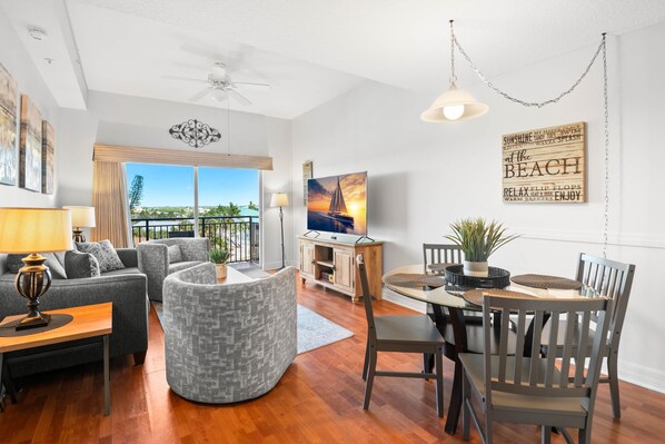A spacious living area with a sofa, armchair, and dining table. A television is mounted on the wall, and there is a large window offering a view of the outdoors.