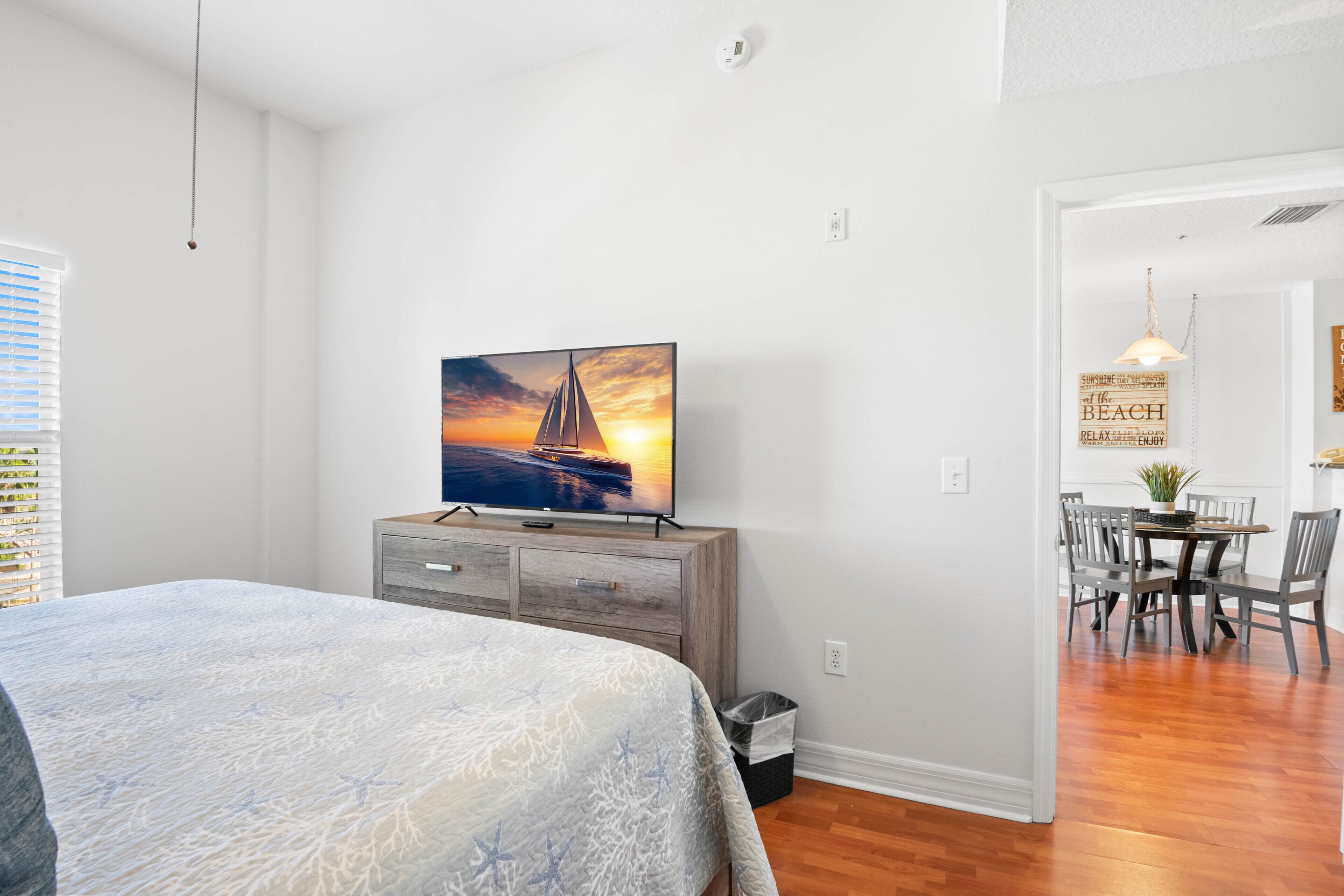 Un dormitorio con cama, un televisor sobre una cómoda de madera, una zona de comedor con mesa y sillas, y un pasillo que conduce a otra habitación.