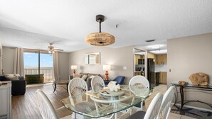 Un salón amplio con una mesa de comedor de vidrio rodeada por sillas de mimbre blanco, un ventilador de techo y vistas al exterior.