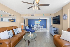 A living room with a brown leather sofa, a glass coffee table, a ceiling fan, a dining area with white chairs, and a TV mounted on the wall.