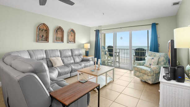 A living room with a grey leather sofa, a wooden coffee table, and a blue armchair. There is a ceiling fan, a TV, and a laptop on a white cabinet. - Sea Breeze 403 | Gulf-Front Balcony Views & Room to Unwind (Madeira Beach)