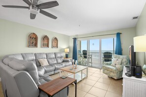 A living room with a grey leather sofa, a wooden coffee table, and a blue armchair. There is a ceiling fan, a TV, and a laptop on a white cabinet. - Sea Breeze 403 | Gulf-Front Balcony Views & Room to Unwind (Madeira Beach)