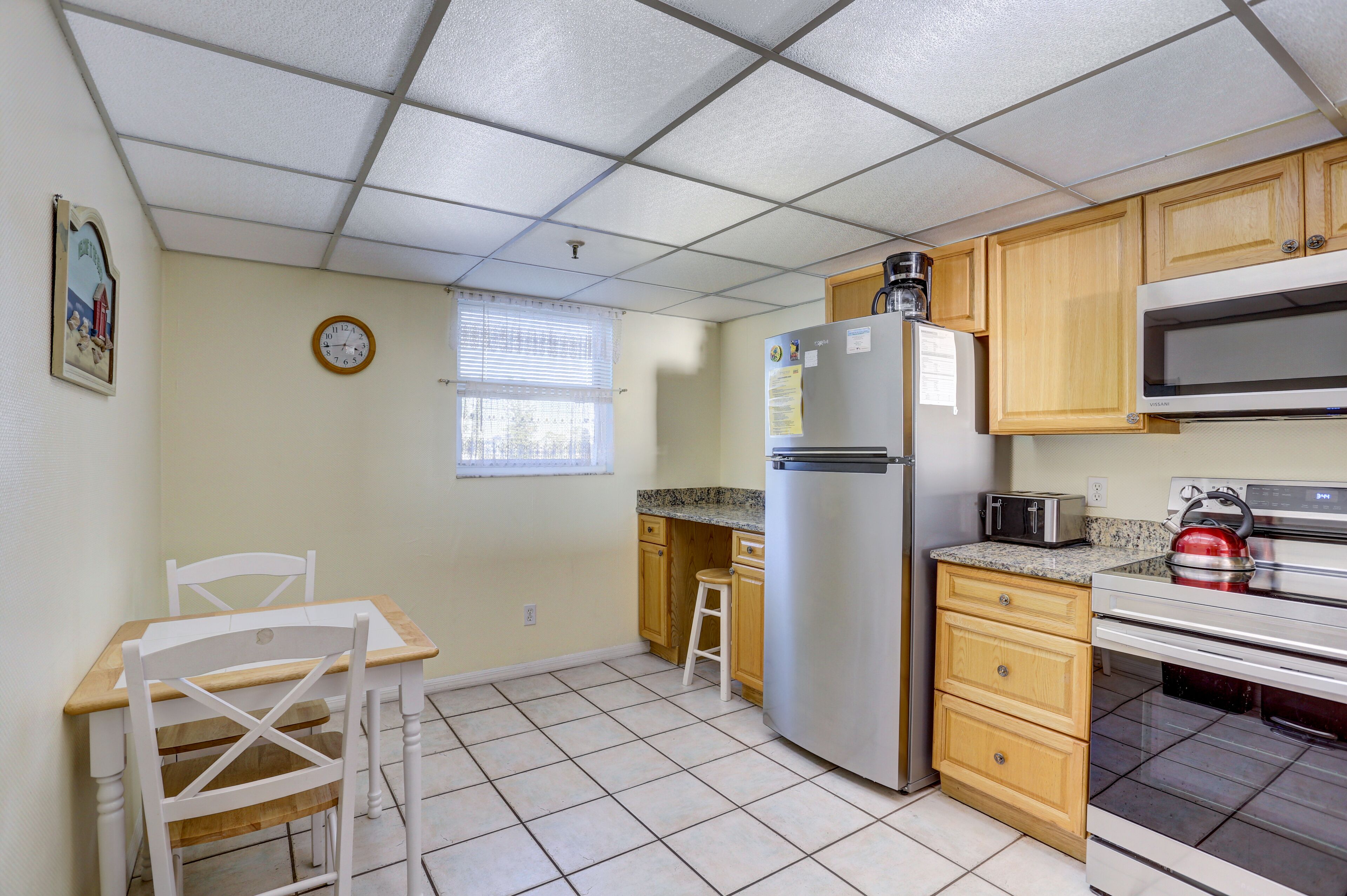 Cocina compacta con refrigerador, microondas y estufa. Incluye una pequeña mesa de comedor con sillas y una ventana con persianas.
