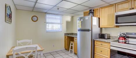 Cocina compacta con refrigerador, microondas y estufa. Incluye una pequeña mesa de comedor con sillas y una ventana con persianas.
