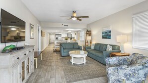 A spacious living room with a ceiling fan, a flat-screen TV, a dining area, and a kitchen in the background.