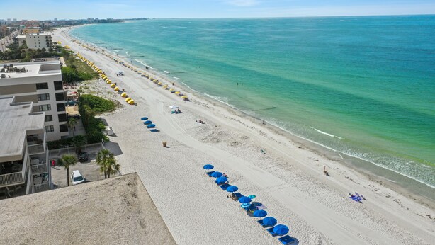 A beach with rows of blue umbrellas and sun loungers, a sandy shoreline, and buildings in the background.