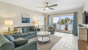 A living room with a sofa, armchairs, a coffee table, and a TV. There is a balcony with a view of palm trees and the ocean.
