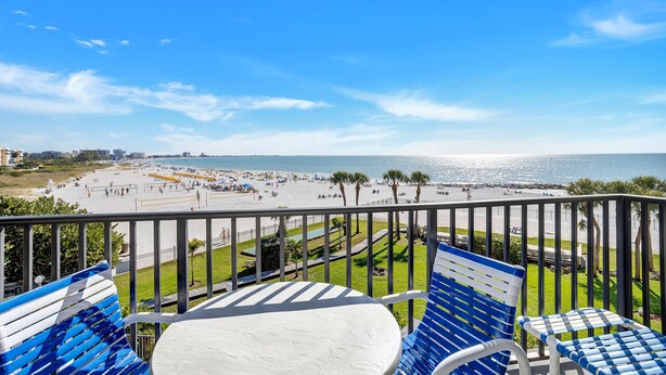 A balcony with blue and white striped chairs and a white table, overlooking a beach with palm trees and a clear blue sky.