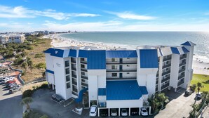 A multi-story hotel with a blue roof and white walls, located near a beach with a parking lot and several cars.