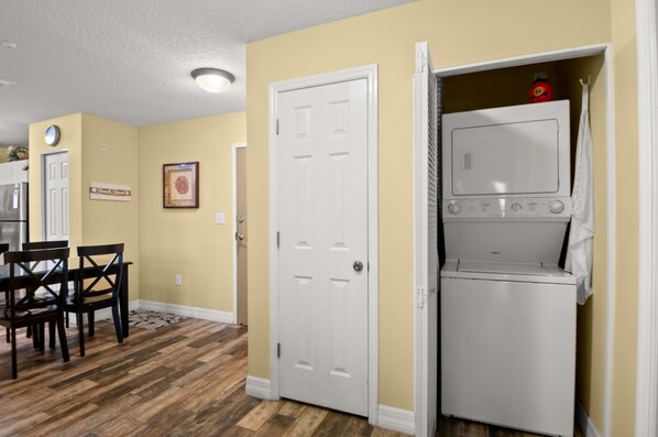 A laundry room with a white washer and dryer, wooden flooring, and a door leading to another room.