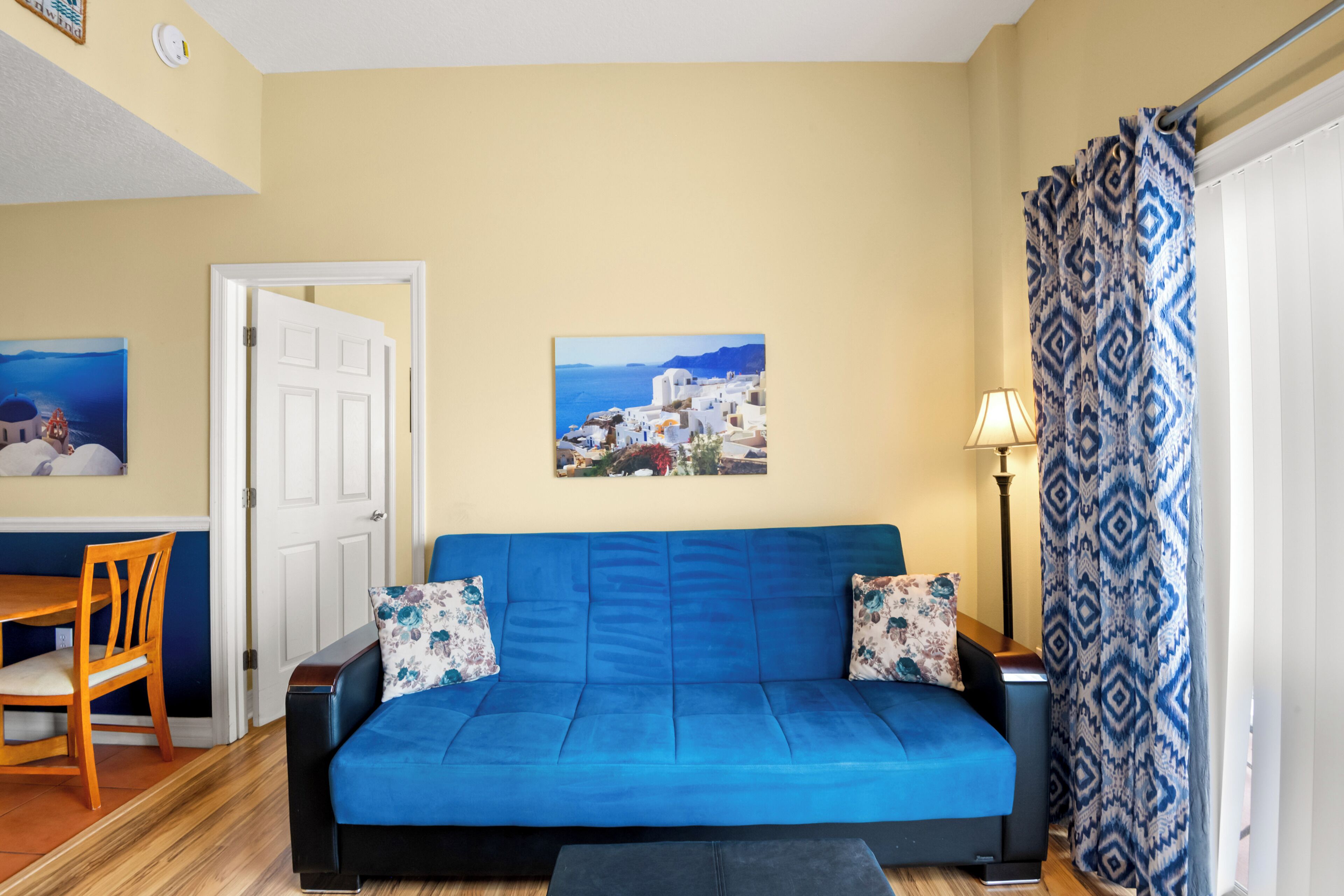 Ein Zimmer mit einem blauen Sofa, einem Esstisch mit Stühlen, einem Gemälde mit einer Küstenansicht und einem gemusterten Vorhang.