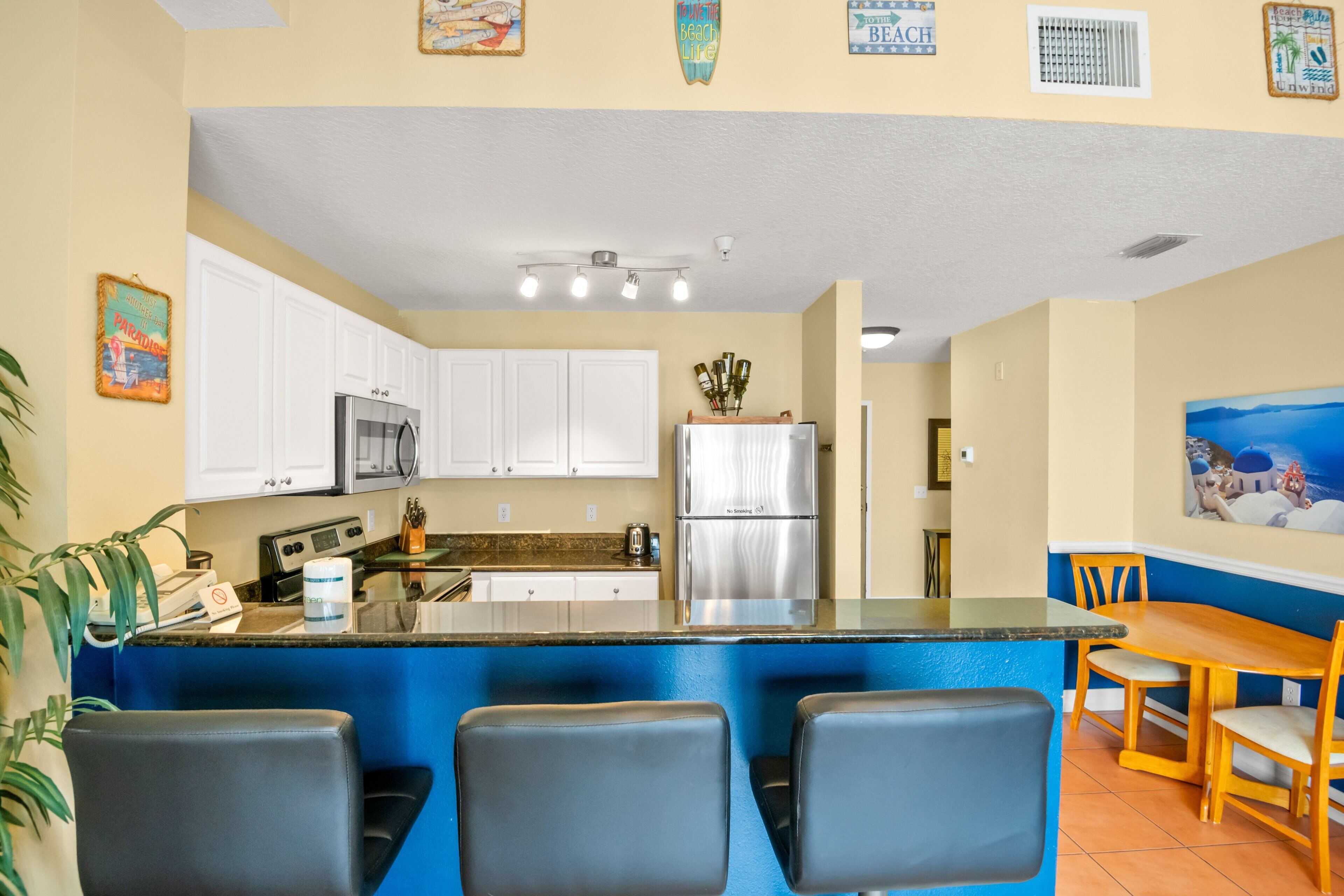 Una cocina con gabinetes blancos, un refrigerador de acero inoxidable, una barra con dos sillas y una mesa de comedor.