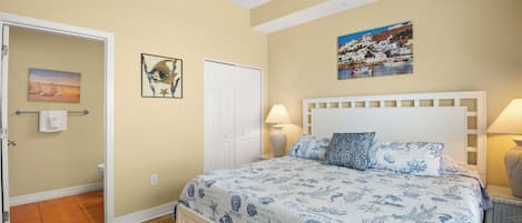 Un dormitorio con una cama, dos lámparas de noche, un cabecero blanco, un cuadro con una escena costera y una puerta que da al baño.