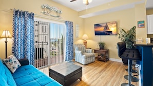 A living room with a blue sofa, a striped armchair, a black ottoman, a ceiling fan, and a painting of a sunset.