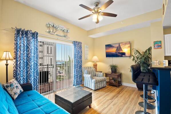 A living room with a blue sofa, a striped armchair, a black ottoman, a ceiling fan, and a painting of a sunset.