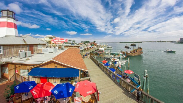 A waterfront pier with a lighthouse, boats, and a restaurant with outdoor seating.