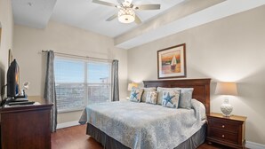 A bedroom with a large bed, a ceiling fan, a window with curtains, a nightstand with a lamp, and a painting on the wall.