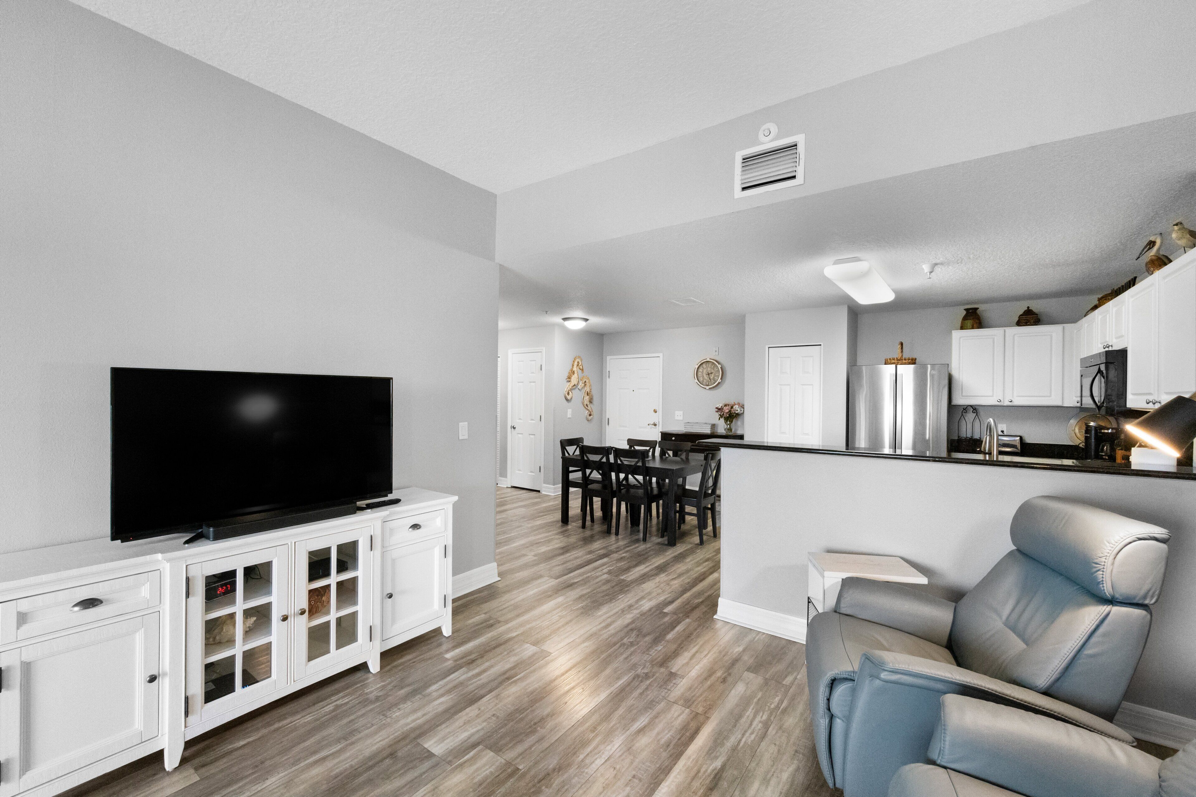 A modern living room with a flat-screen TV, a dining area with a table and chairs, and a kitchen area with cabinets and appliances.