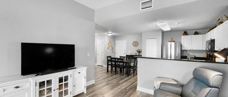 A modern living room with a flat-screen TV, a dining area with a table and chairs, and a kitchen area with cabinets and appliances.