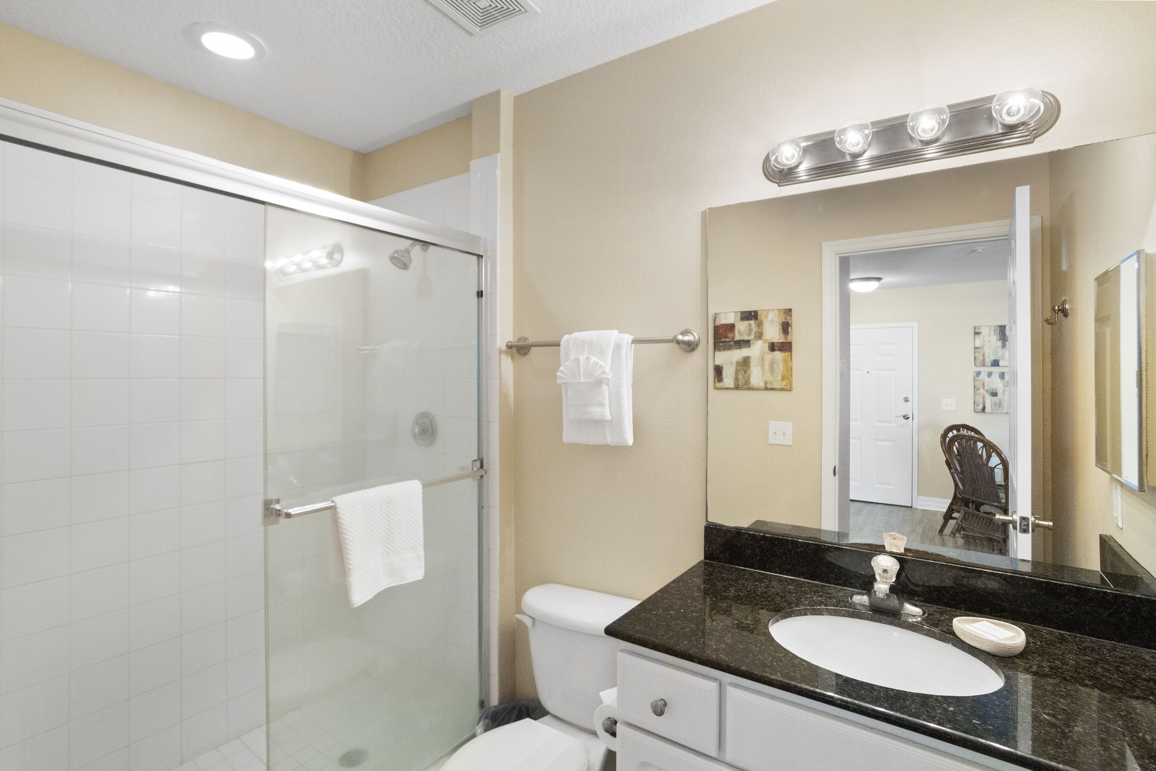 A bathroom with a glass shower enclosure, a white toilet, a black countertop sink, and a mirror with lights above it.