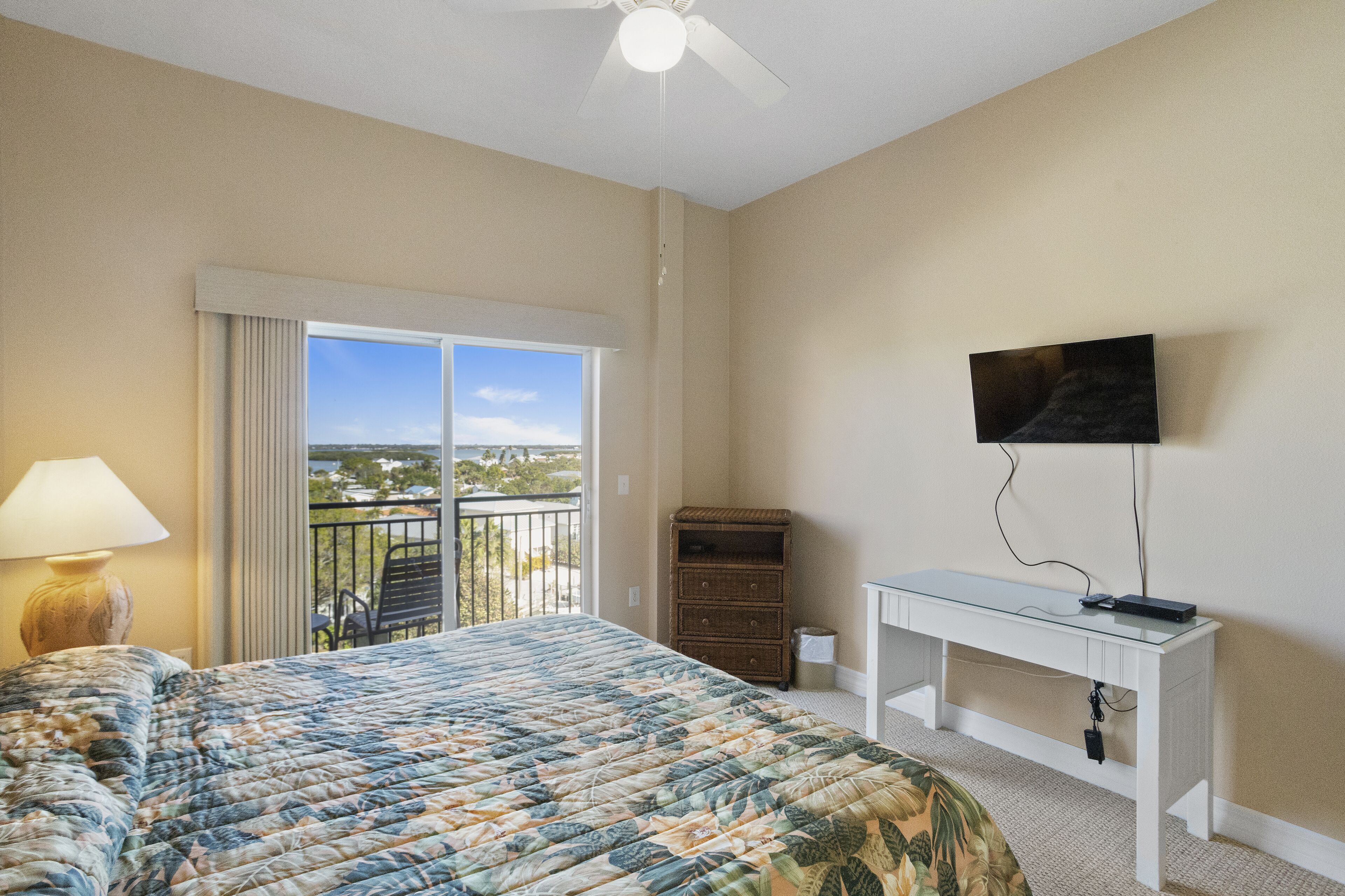 A bedroom with a bed, a desk, a TV, a balcony with a view, and a lamp.