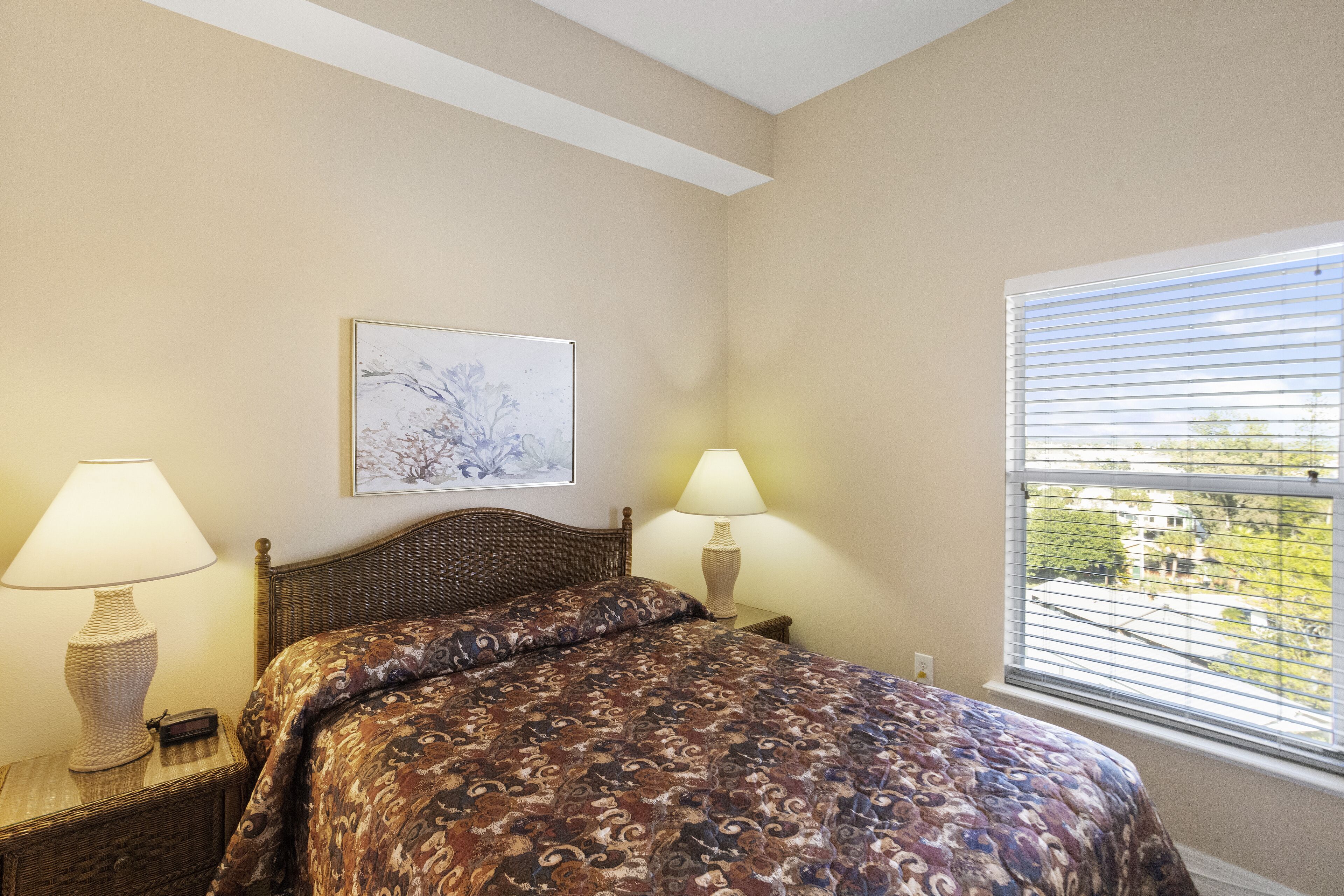 A bedroom with a bed, two bedside lamps, a nightstand, a framed picture on the wall, and a window with blinds.