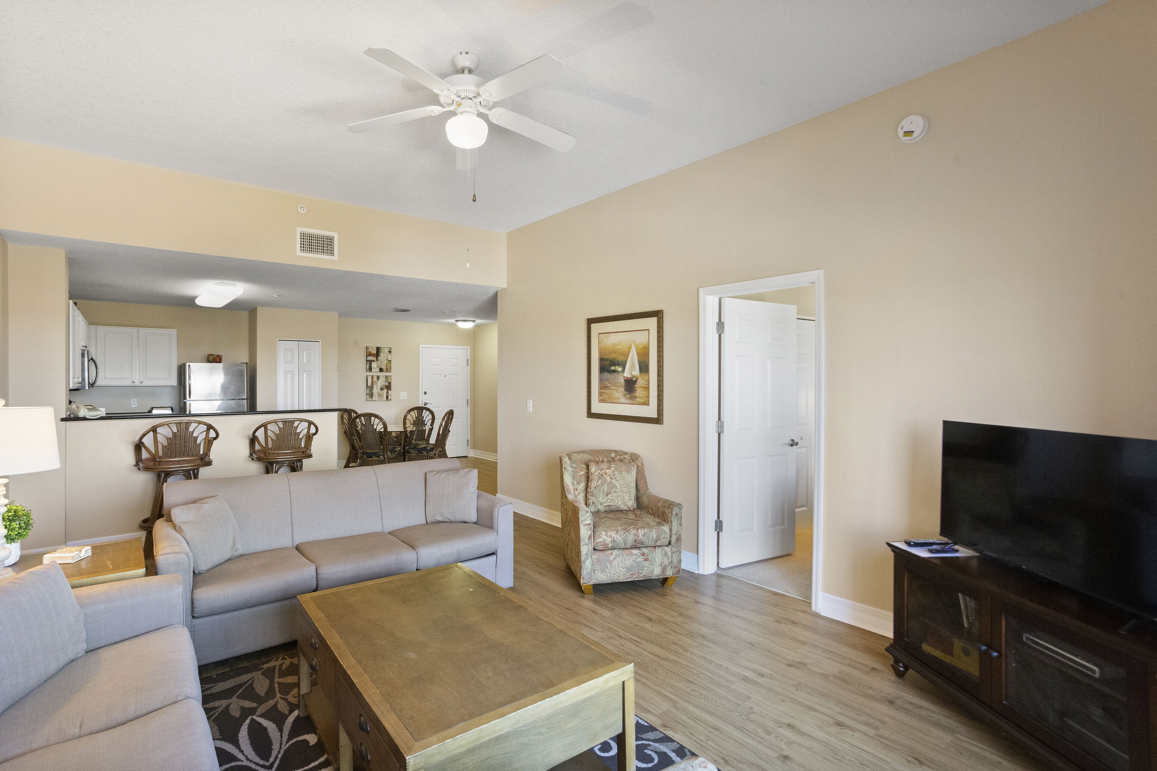 A living room with a sofa, armchair, coffee table, TV, and a kitchen area in the background.