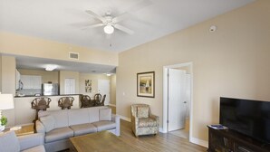 A living room with a sofa, armchair, coffee table, TV, and a kitchen area in the background.