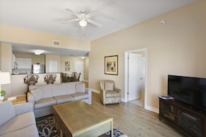 A living room with a sofa, armchair, coffee table, TV, and a kitchen area in the background.