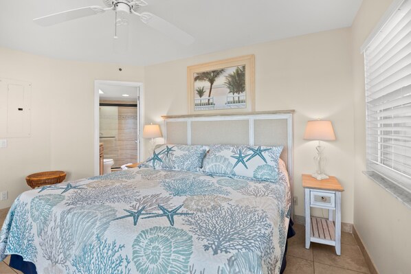 A bedroom with a bed, two bedside tables, a ceiling fan, a window with blinds, and a door leading to a bathroom. - 🌅 Prime View Condo — Gulf Strand Resort, St. Pete Beach (St Pete Beach)