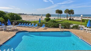 A swimming pool with blue chairs and umbrellas, a view of the beach, and a clear sky.