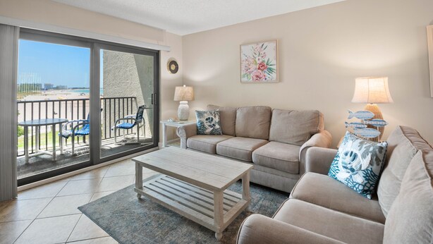 A living room with a sofa, coffee table, and two lamps. There is a balcony with chairs and a view of the beach.