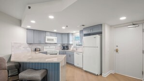 A modern kitchen with grey cabinets, a white refrigerator, a microwave, and a built-in island with bar stools. - 🌴 Gulf View Condo — Gulf Strand Resort, St. Pete Beach (St Pete Beach)