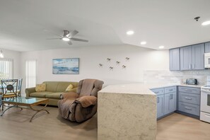 Uma sala moderna com um sofá, uma poltrona reclinável, uma mesa de centro de vidro, um ventilador de teto e uma área de cozinha com armários e um micro-ondas.